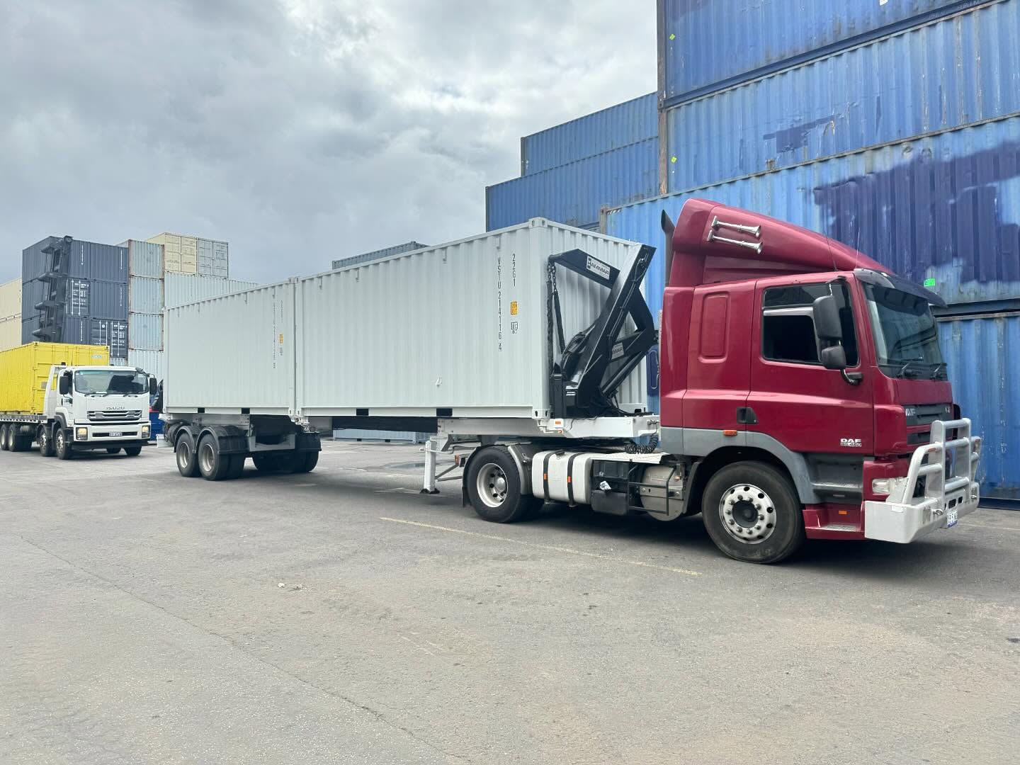 Red DAF side loader truck carrying white shipping containers in a container yard
