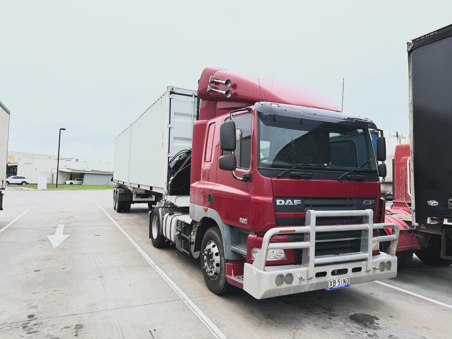 Red DAF prime mover with a white container on its side loader trailer in a truck car park