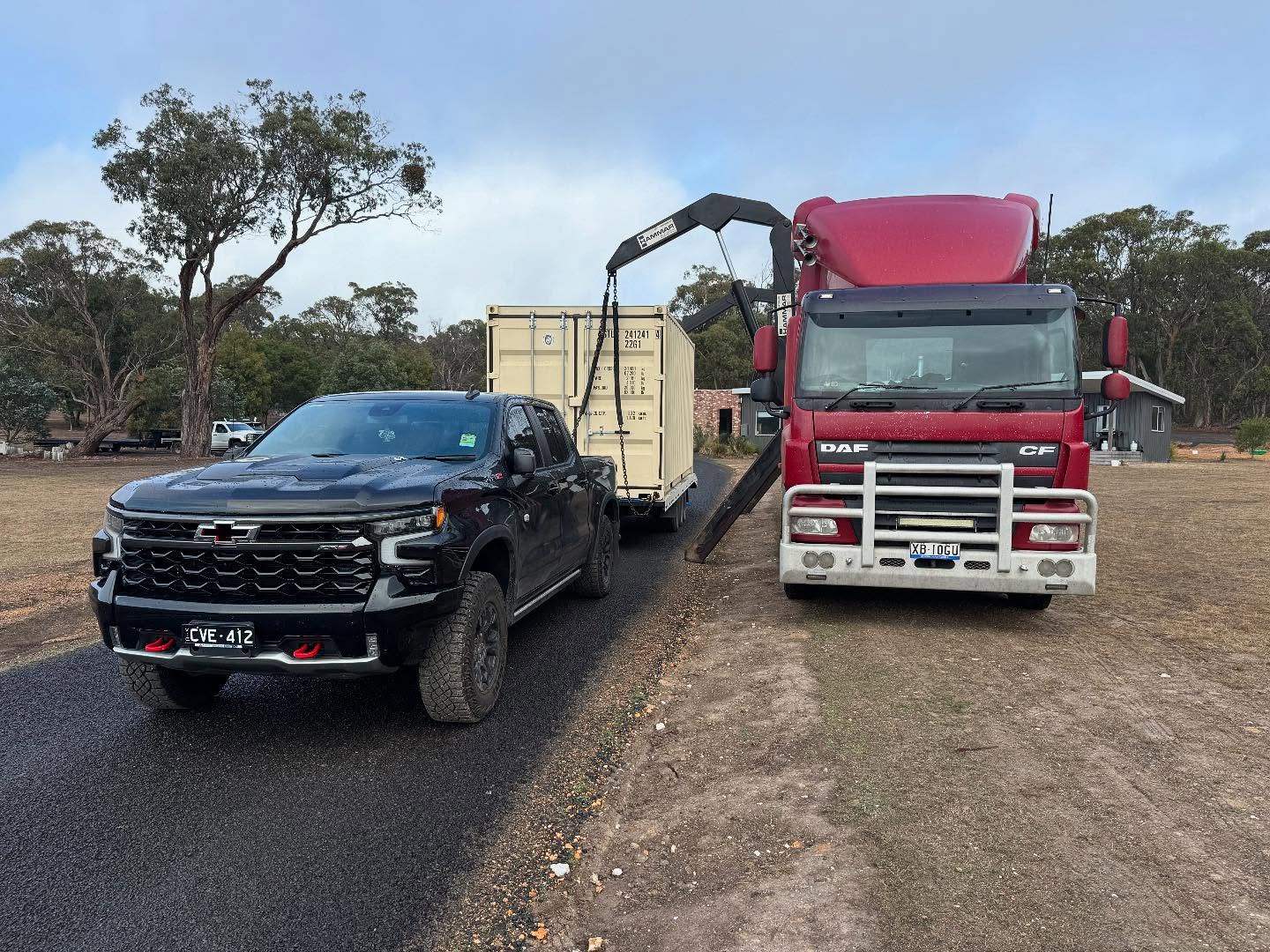 Red DAF side loader truck holding a shipping container on its lifting arms beside a black Chevrolet ute
